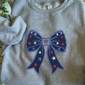 May include: A grey sweatshirt with a navy blue embroidered bow design. The bow features red, white, and blue star details. The bow's centre has a heart design with red and blue stripes. The sweatshirt has a crew neck and long sleeves.