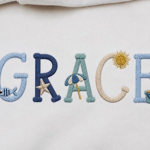 May include: Embroidered word "GRACE" in various shades of blue, beige, and green. Each letter is decorated with beach-themed elements like a fish, starfish, umbrella, sun, and sand pail. The design is on a white fabric.