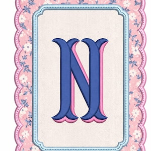 Puede incluir: Un parche rectangular bordado con un borde festoneado con un patrón floral rosa. El centro es un rectángulo de color crema con un borde azul claro, que contiene una letra N grande, negrita y azul oscuro con un efecto de sombra rosa.