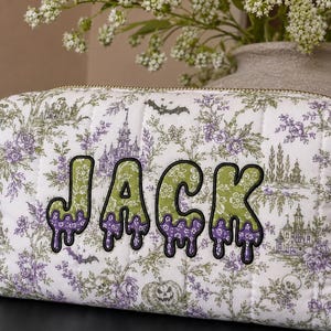 May include: A rectangular quilted fabric pouch featuring a white background with a repeating pattern of purple and green haunted castles, bats, and pumpkins. The name JACK is appliqued in large, green and purple dripping-style letters.
