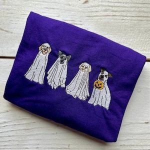 以下が含まれることがあります： 紫色のTシャツに、ハロウィンのために幽霊に扮した4匹の犬の刺繍デザインが施されています。犬たちは白いシーツを着て、頭が顔を出しています。1匹の犬が小さなオレンジ色のカボチャを持っています。