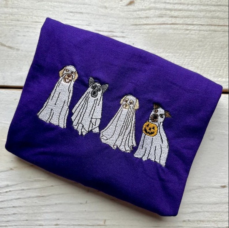以下が含まれることがあります： 紫色のTシャツに、ハロウィンのために幽霊に扮した4匹の犬の刺繍デザインが施されています。犬たちは白いシーツを着て、頭が顔を出しています。1匹の犬が小さなオレンジ色のカボチャを持っています。