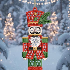 Puede incluir: Adorno navideño bordado de un soldado cascanueces. El adorno es rojo, verde y dorado con un bigote negro y una ramita de hojas verdes. Cuelga de un hilo dorado sobre un fondo nevado borroso.