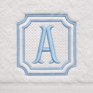 May include: A white terry cloth towel featuring a central embroidered square emblem with scalloped corners. The emblem has a textured white background, a light blue border, and a large, stylized light blue letter A in the center.