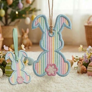 May include: Two Easter bunny-shaped ornaments with pastel rainbow stripes and a light blue scalloped border. One ornament is hanging by a twine string, and the other has a light green ribbon. A pink flower is at the bottom.