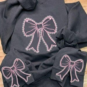 May include: Dark gray hoodie with a large pink bow design on the front and smaller bow designs on the sleeves. The bows have a lace-like trim. The hoodie is made of a soft material.