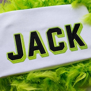 May include: White fabric with the name "JACK" embroidered in black letters with a neon green outline. The fabric is surrounded by bright green feathers.