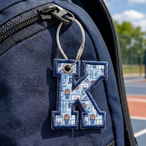 May include: A letter K keychain featuring a light blue fabric with a repeating basketball-themed pattern, including basketballs, hoops, and sneakers. The letter has a dark blue stitched border and is attached to a silver metal cable ring.