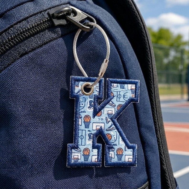 May include: A letter K keychain featuring a light blue fabric with a repeating basketball-themed pattern, including basketballs, hoops, and sneakers. The letter has a dark blue stitched border and is attached to a silver metal cable ring.