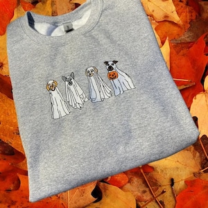 以下が含まれることがあります： ハロウィン用のゴーストに扮した4匹の犬が刺繍されたグレーのスウェットシャツ。犬たちは白いゴーストの衣装を着て、頭が顔を出しています。1匹の犬が小さなオレンジ色のカボチャを持っています。