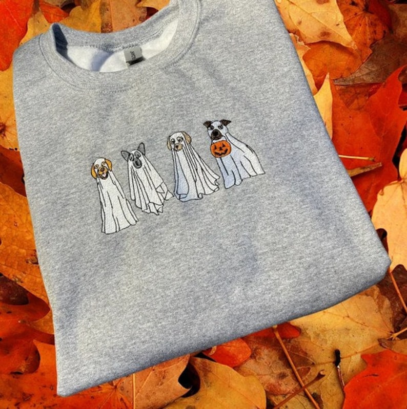 以下が含まれることがあります： ハロウィン用のゴーストに扮した4匹の犬が刺繍されたグレーのスウェットシャツ。犬たちは白いゴーストの衣装を着て、頭が顔を出しています。1匹の犬が小さなオレンジ色のカボチャを持っています。