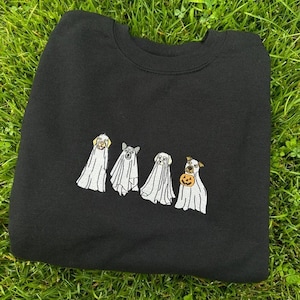 以下が含まれることがあります： ハロウィンのために幽霊に扮した4匹の犬の刺繍が施された黒いスウェットシャツ。1匹の犬がカボチャを持っています。