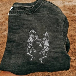 Puede incluir: Sudadera gris oscuro con un dise&ntilde;o bordado en blanco de dos esqueletos bailando. La sudadera de cuello redondo est&aacute; doblada, mostrando el gr&aacute;fico en la parte delantera. La tela parece suave y c&oacute;moda.