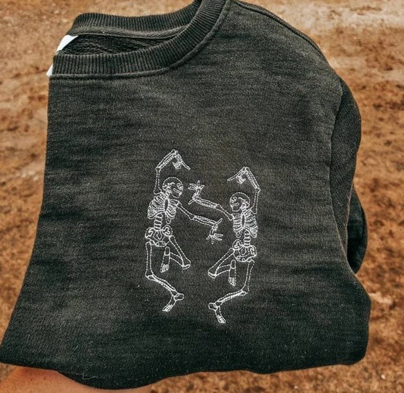 Puede incluir: Sudadera gris oscuro con un dise&ntilde;o bordado en blanco de dos esqueletos bailando. La sudadera de cuello redondo est&aacute; doblada, mostrando el gr&aacute;fico en la parte delantera. La tela parece suave y c&oacute;moda.