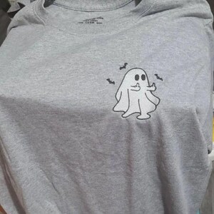May include: A gray t-shirt featuring a cartoon ghost giving the middle finger, with three small bat illustrations. The ghost is white with black eyes and a black outline. The shirt is made of a soft material.