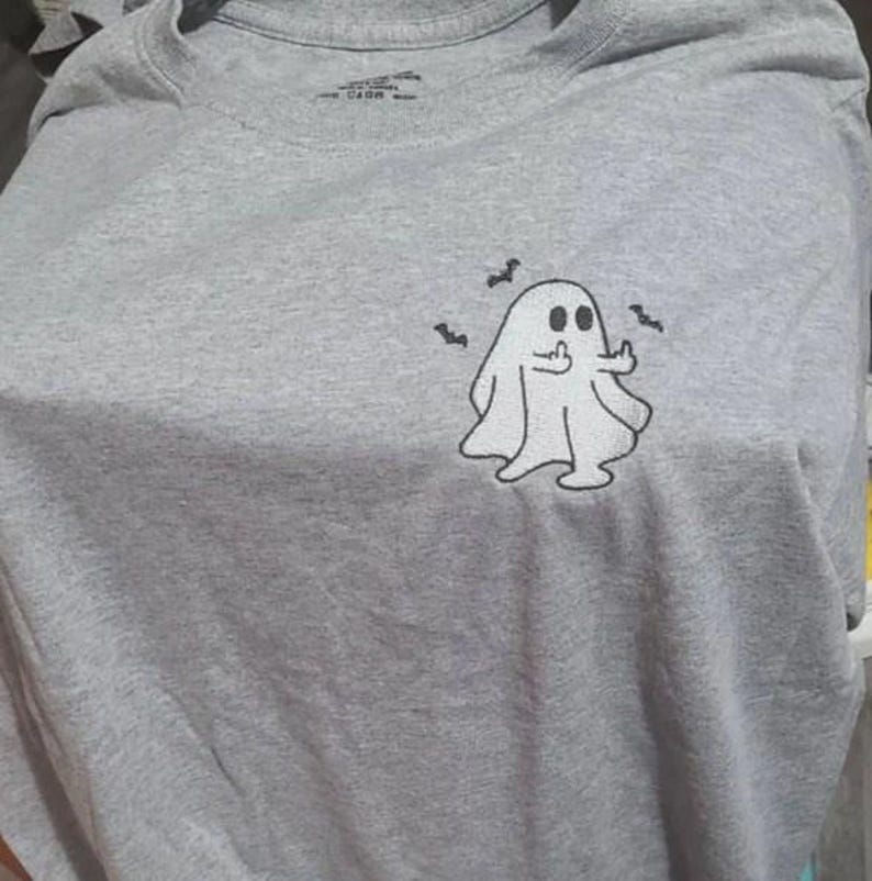 May include: A gray t-shirt featuring a cartoon ghost giving the middle finger, with three small bat illustrations. The ghost is white with black eyes and a black outline. The shirt is made of a soft material.