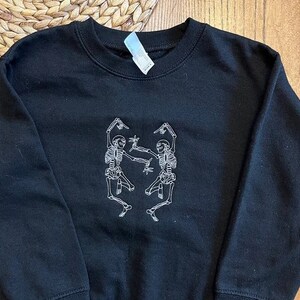 Puede incluir: Sudadera negra de cuello redondo con un dise&ntilde;o bordado en blanco de dos esqueletos bailando. Los esqueletos est&aacute;n representados en una pose juguetona, con los brazos levantados. La sudadera es de material suave.