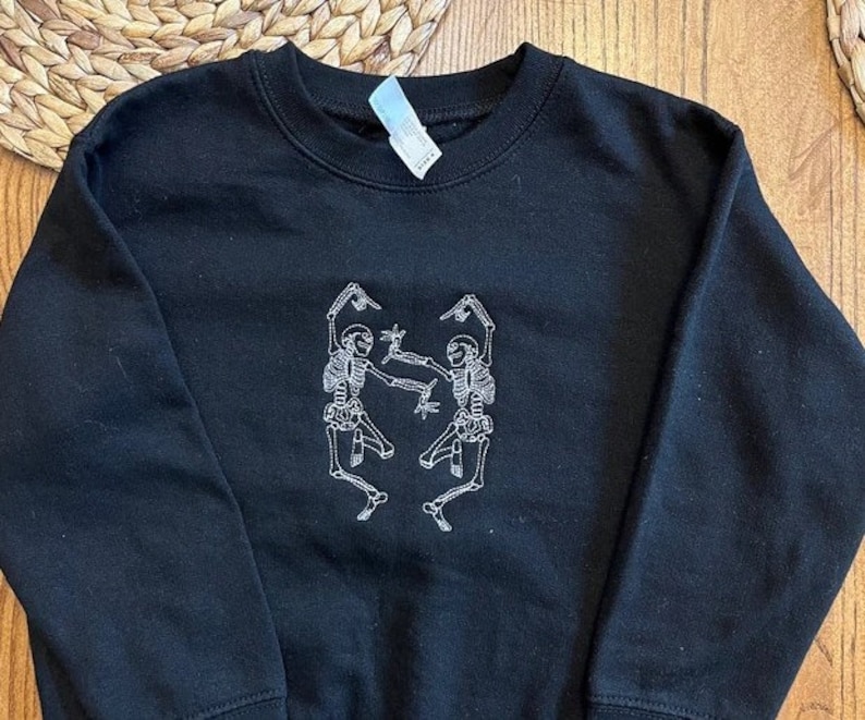 Puede incluir: Sudadera negra de cuello redondo con un dise&ntilde;o bordado en blanco de dos esqueletos bailando. Los esqueletos est&aacute;n representados en una pose juguetona, con los brazos levantados. La sudadera es de material suave.