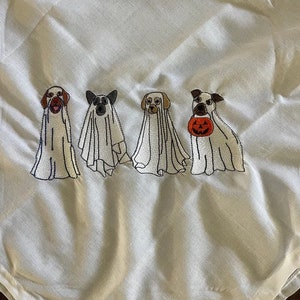 以下が含まれることがあります： ハロウィンのために幽霊に扮した刺繍された犬が5匹います。犬たちは一列に並んでおり、右側の犬は小さなオレンジ色のカボチャを持っています。