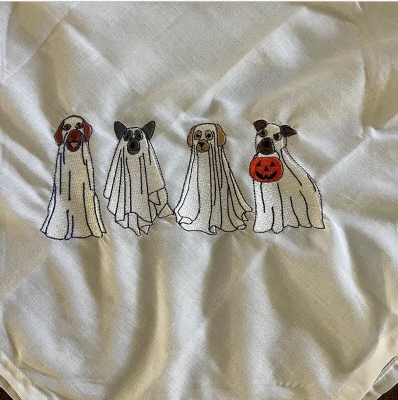 以下が含まれることがあります： ハロウィンのために幽霊に扮した刺繍された犬が5匹います。犬たちは一列に並んでおり、右側の犬は小さなオレンジ色のカボチャを持っています。