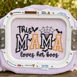 Op de afbeelding: Een lichtpaarse stof in een witte borduurring met zwarte en oranje geborduurde tekst die This MAMA loves her boos leest, vergezeld door zwarte vleermuissilhouetten en een spinnenwebontwerp boven de belettering.