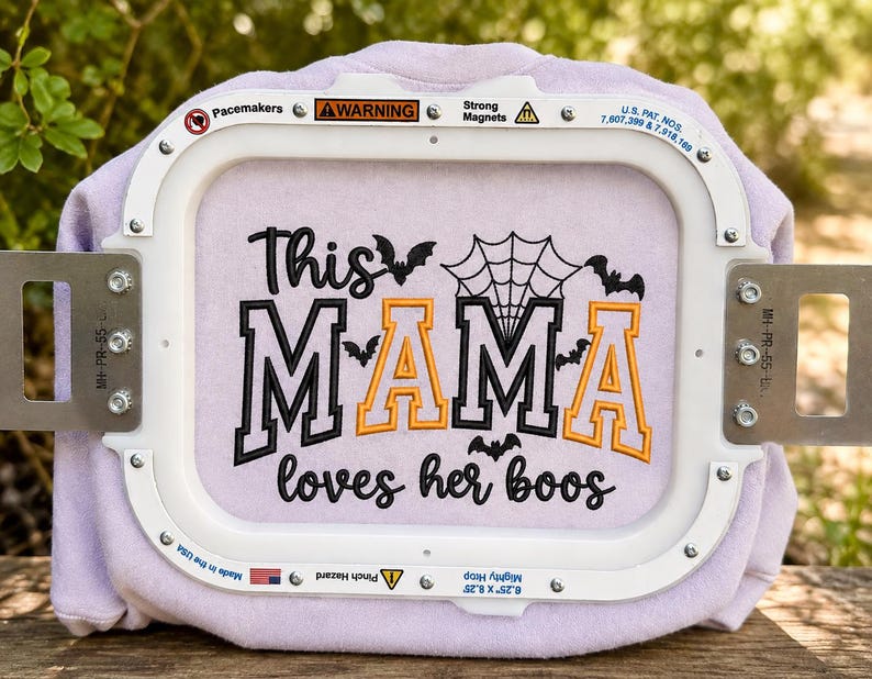 Op de afbeelding: Een lichtpaarse stof in een witte borduurring met zwarte en oranje geborduurde tekst die This MAMA loves her boos leest, vergezeld door zwarte vleermuissilhouetten en een spinnenwebontwerp boven de belettering.