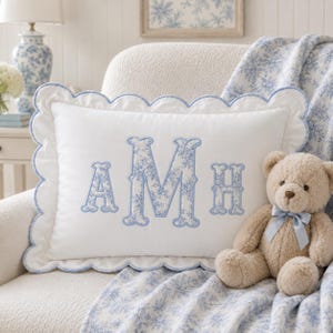 May include: A rectangular white pillow with a scalloped edge trimmed in light blue. The center features the embroidered monogram letters AMH in a blue and white floral pattern. A light brown teddy bear with a light blue ribbon sits nearby.