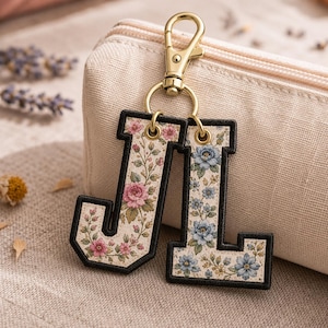 May include: A keychain featuring two embroidered letter charms, J and L, attached to a gold-toned metal lobster clasp. The letter J is decorated with pink floral patterns, while the letter L features blue floral designs, both with black borders.