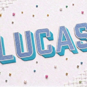 May include: The name LUCAS is embroidered in a varsity-style font with light blue letters featuring a dark blue outline and a bright turquoise drop shadow. The embroidery is set against a white fabric background scattered with small colourful beads.