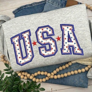 Op de afbeelding: Een grijze sweatshirt met het geborduurde woord "USA". De letters zijn blauw omlijnd en gevuld met rode en witte sterren. Rode sterren zijn tussen de letters geplaatst. De sweatshirt is op een blauwe jeans gevouwen.