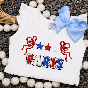 May include: A white sleeveless top with ruffled shoulders and hem, featuring an embroidered design of two red bows, three stars in blue, red, and white, and the text PARIS in red, white, and blue. A light blue ribbon bow is attached at the shoulder.