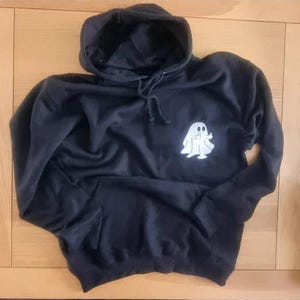 May include: Black hooded sweatshirt with a white ghost graphic on the front. The sweatshirt has a drawstring hood and a front pocket. The ghost graphic is a simple, cartoon-like design.