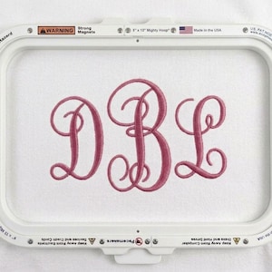 Puede incluir: Un bastidor de bordado blanco con las letras "DBL" en un color rosa oscuro y fuente cursiva. El bastidor tiene advertencias y el texto "Made in the USA".