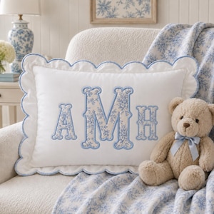 May include: A rectangular white throw pillow with a scalloped edge trimmed in light blue. The centre features the embroidered letters AMH in a blue and white floral pattern. A light brown teddy bear with a light blue ribbon sits beside it.