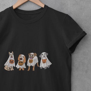 Puede incluir: Camiseta negra con un gr&aacute;fico de cuatro perros disfrazados de fantasmas que llevan bolsas de truco o trato. Los perros llevan disfraces de fantasmas blancos con cubos de calabaza naranja y negro.