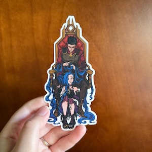May include: A sticker featuring a cartoon illustration of two characters seated on a throne. One character is seated on the throne, and the other is seated in front of the first character. The seated character has long blue hair. The sticker is held by a hand.