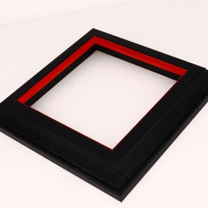 May include: A black picture frame with a red inner border. The frame has a textured wood-like finish.