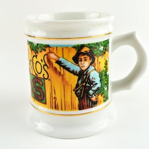 May include: White ceramic mug with a decorative illustration. The mug features a boy in a hat leaning against a wooden fence. The mug has a gold trim and a handle. The text on the mug reads "S.CO'S".