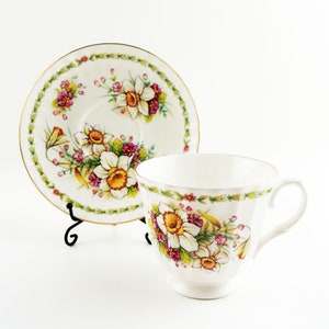 May include: White teacup and saucer set with a floral daffodil design. The cup and saucer have a gold trim and a black metal stand. The floral pattern includes white daffodils with yellow centers, pink and purple flowers, and green leaves.