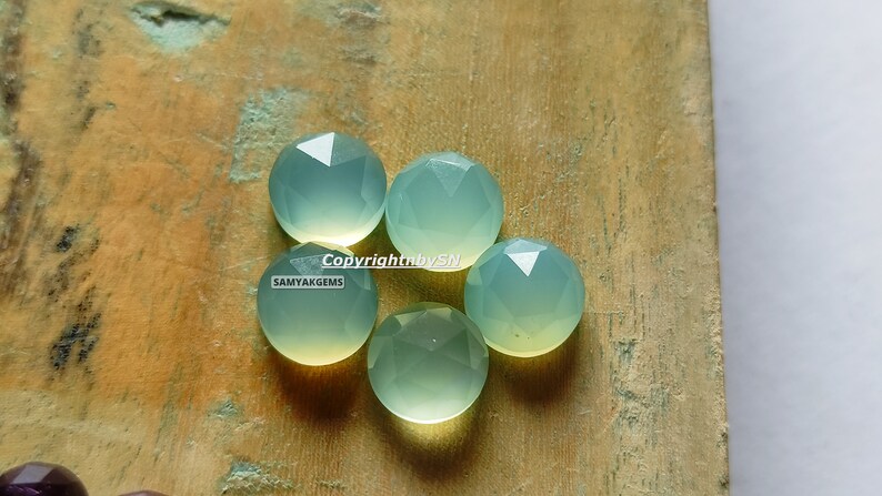 May include: Five faceted, light green chalcedony gemstones on a wooden surface. Copyright by SN SAMYAKGEMS.