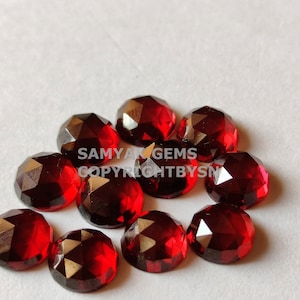 May include: A group of ten faceted red gemstones, likely garnets, arranged on a white surface.  The gemstones are all approximately the same size and shape, with a slightly rounded oval form.  The gemstones have a deep red color and a high shine.