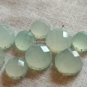 May include: A group of ten light green, faceted, round gemstone cabochons. The cabochons are arranged in a cluster on a white fabric background.