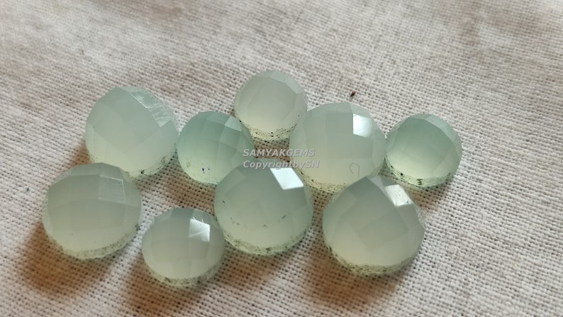 May include: A group of ten light green, faceted, round gemstone cabochons. The cabochons are arranged in a cluster on a white fabric background.