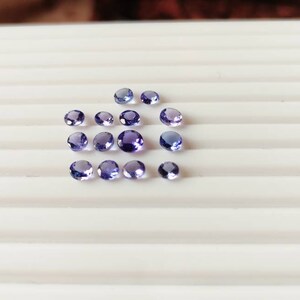 May include: A collection of 17 small, round, purple gemstones arranged in a loose pattern on a white surface.