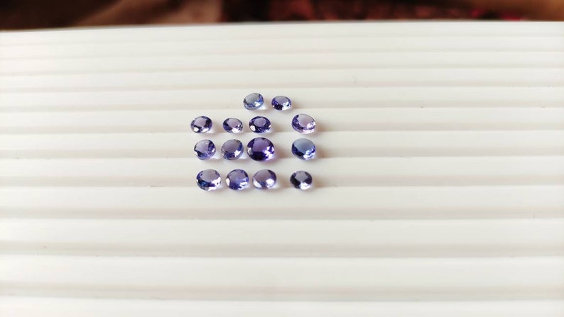 May include: A collection of 17 small, round, purple gemstones arranged in a loose pattern on a white surface.