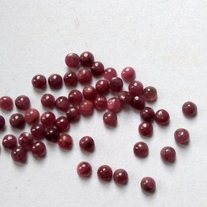 Puede incluir: Un conjunto de pequeñas gemas redondas, de color rojo oscuro, dispersas sobre una superficie blanca. Las gemas pulidas tienen un aspecto brillante, con ligeras variaciones de color. Podrían utilizarse para la fabricación de joyas u otras manualidades.
