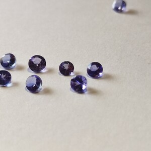May include: A group of eight round, purple gemstones on a white surface.