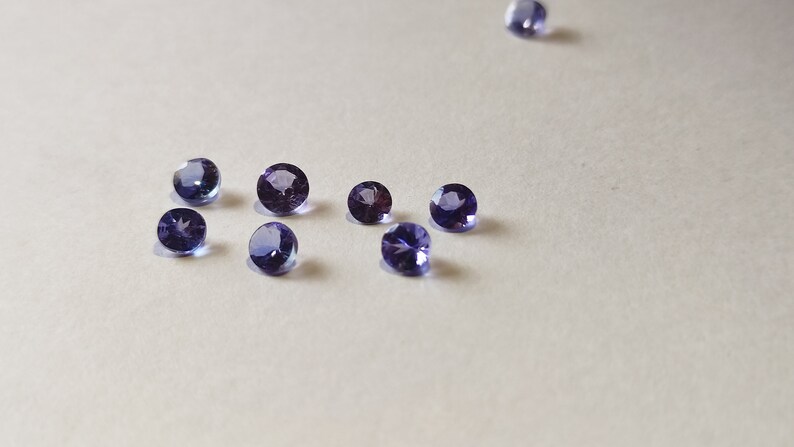 May include: A group of eight round, purple gemstones on a white surface.