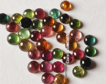 May include: A collection of polished, round gemstones in various colors, including green, red, orange, and black. The gems are scattered on a white surface, with a copyright notice visible. These gemstones are likely used for jewelry making.