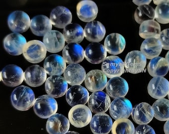 Puede incluir: Una colección de cabujones redondos de piedra lunar pulida. Las piedras muestran una base blanca lechosa con destellos iridiscentes azules y amarillos. Las gemas están dispersas sobre una superficie oscura, mostrando su belleza natural y potencial para la joyería.